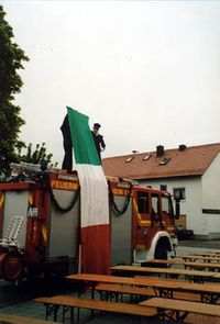 Zwei Feuerwehrler stehen auf dem Dach des LF16 und halten eine Italienflagge, die bis zum Boden reicht